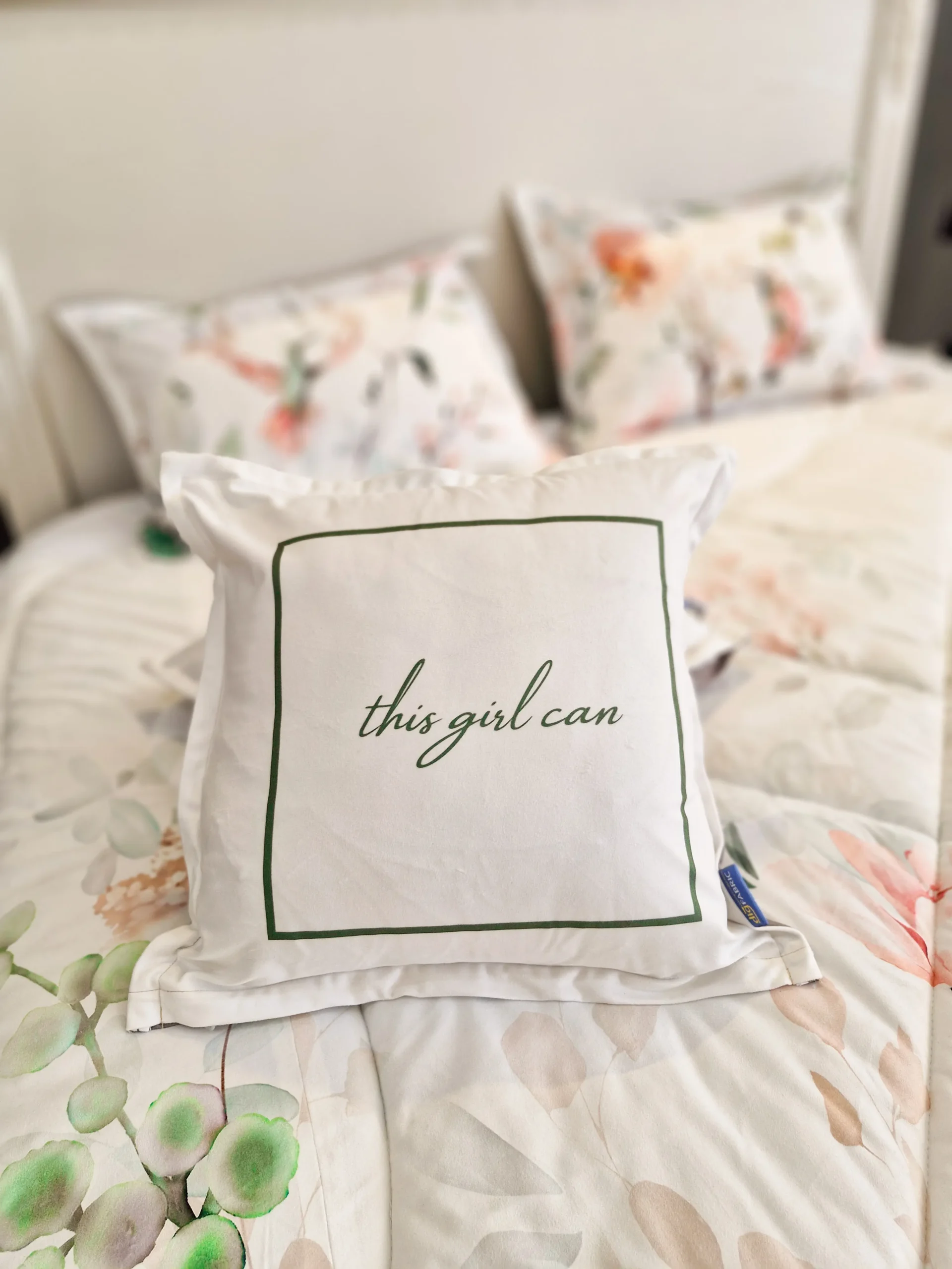 نمای نزدیک کوسن سفید چاپی طرح «this girl can» روی روتختی ولارا با زمینه گل‌بهاری